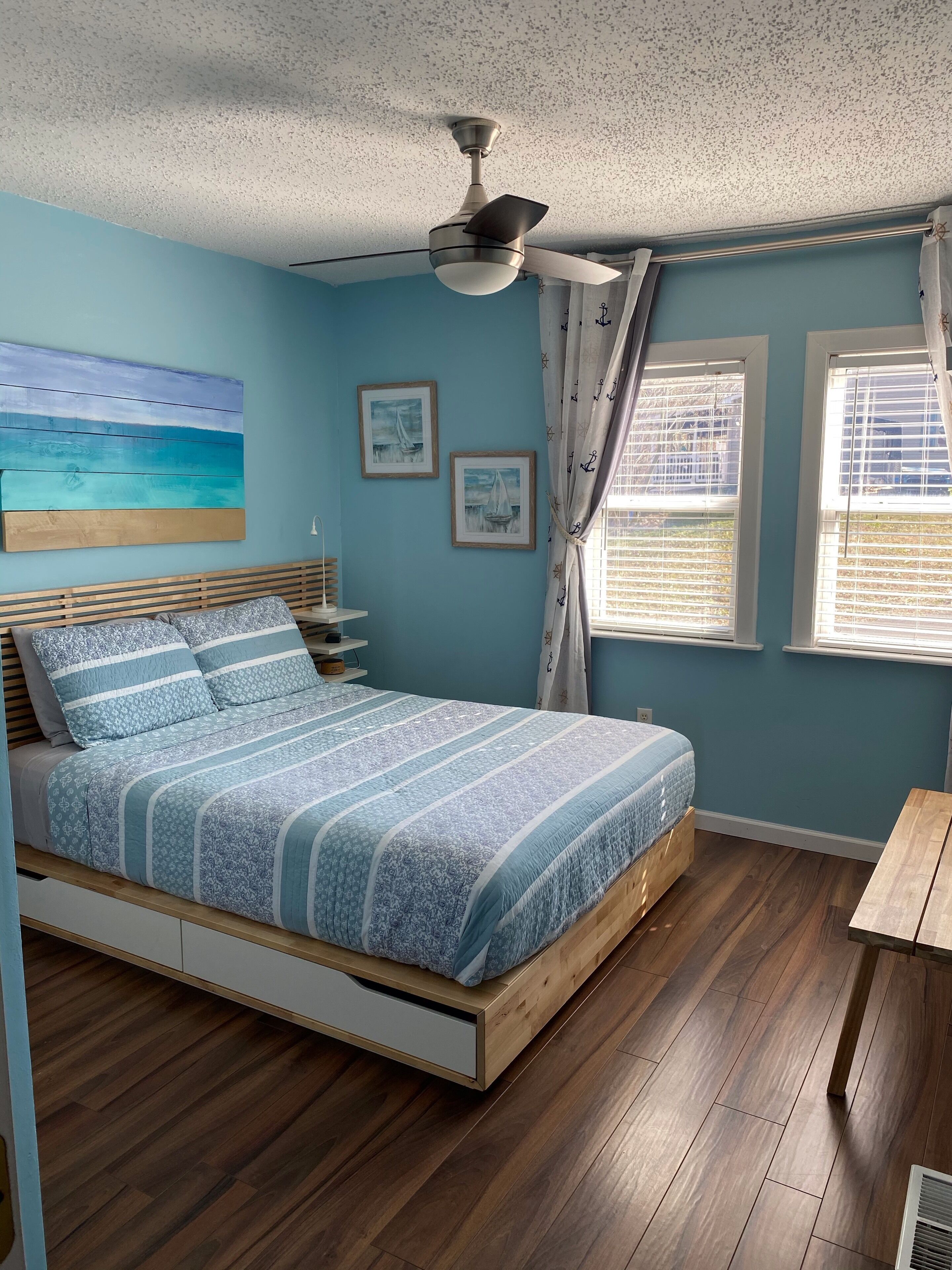 Un dormitorio con cama, ventilador de techo, dos ventanas con cortinas y un cuadro en la pared.