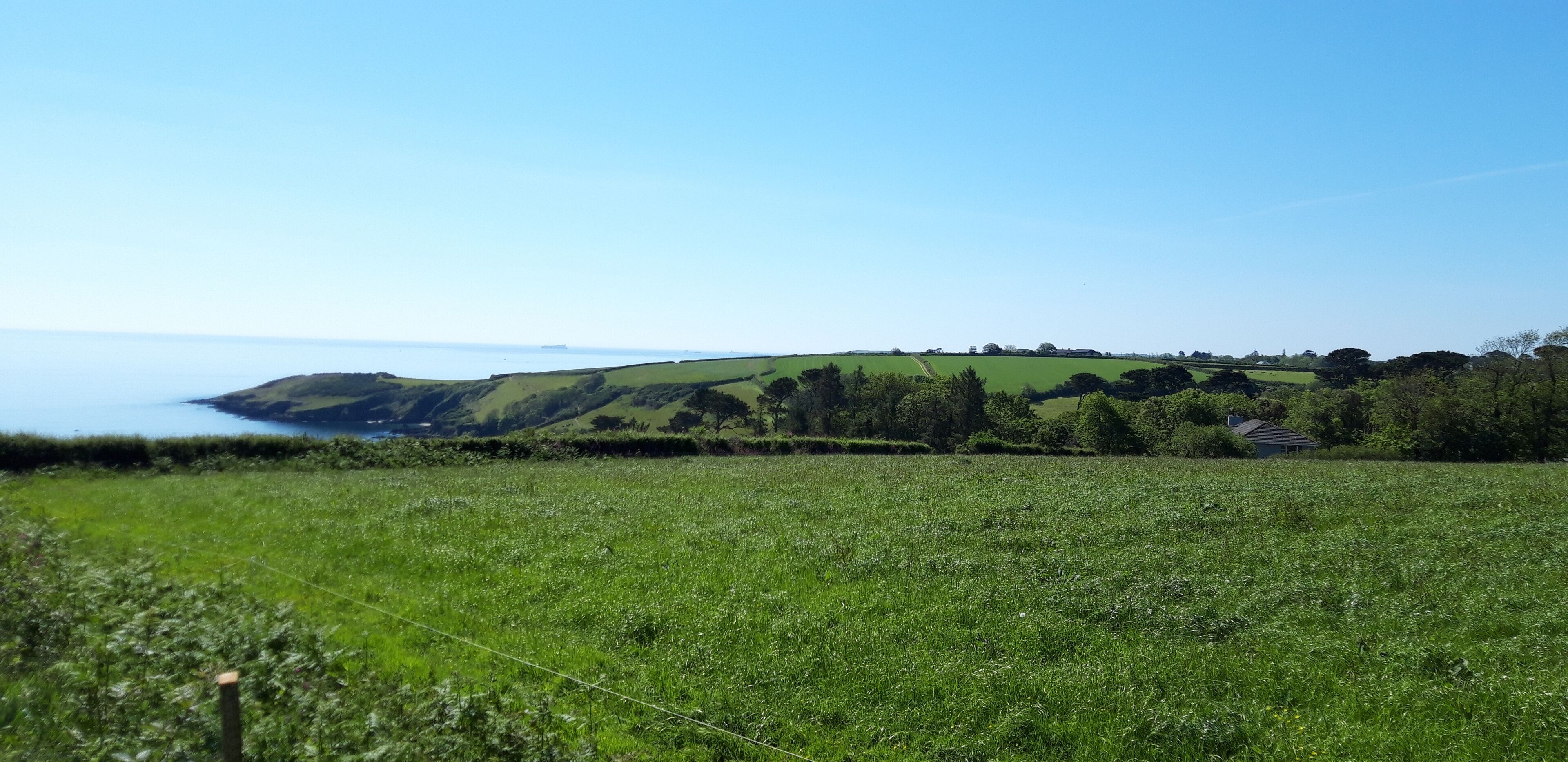 Paisaje rural con un campo, una casa pequeña y una vista distante del mar.