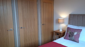A bedroom with a bed, a nightstand with a lamp, and wooden wardrobes. - Trelawne Garden Annexe (Falmouth)