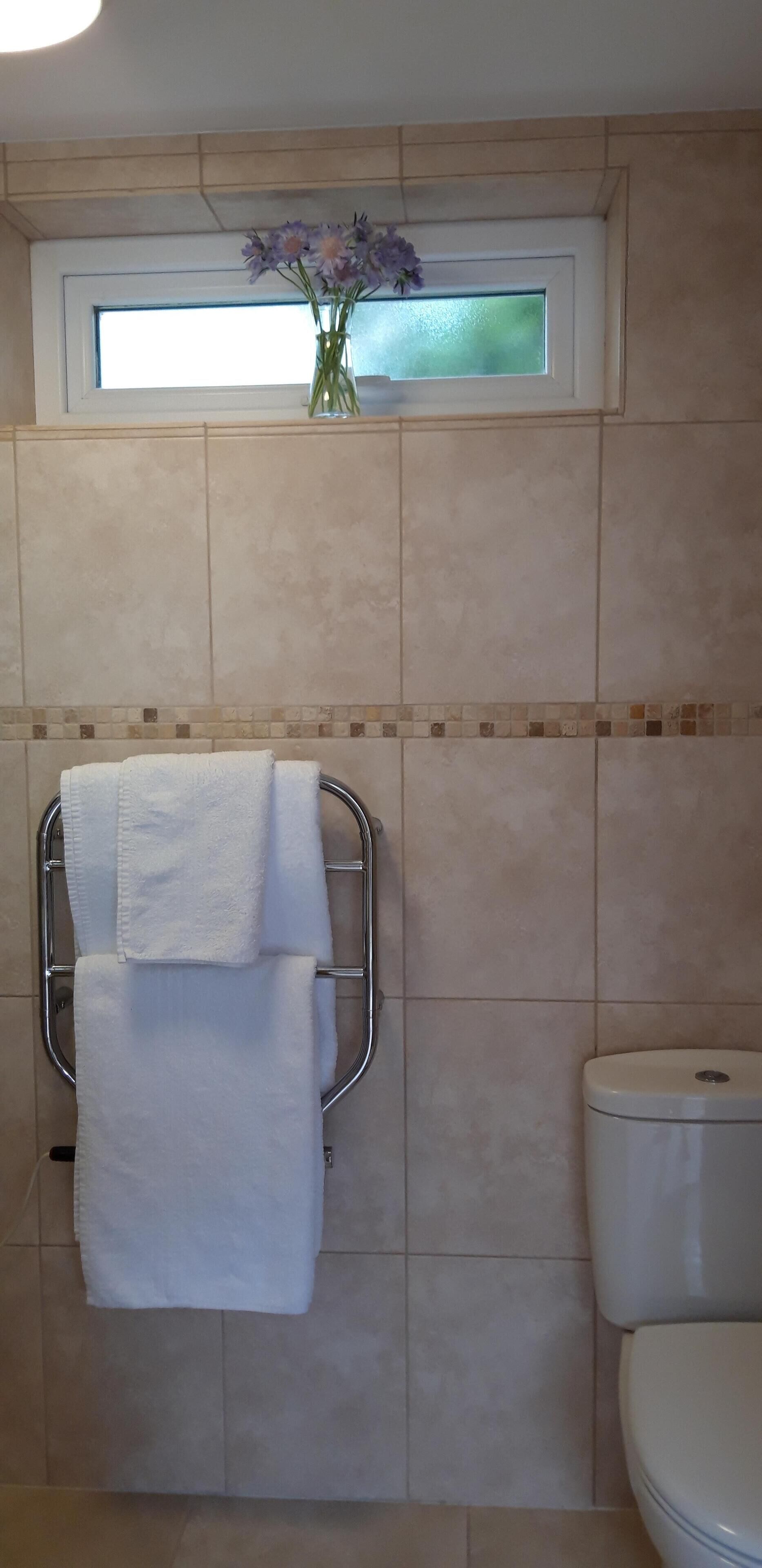 Un baño con baldosas color crema, una ventana con vistas, un inodoro blanco y un perchero con dos toallas.