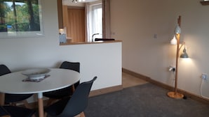A dining area with a round table, black chairs, a white wall, a wooden counter, and a floor lamp. - Trelawne Garden Annexe (Falmouth)