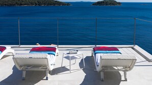 A balcony with two white wicker lounge chairs, each with a striped blanket, overlooking a clear blue sea with distant islands. - Villa Desire (Blato)