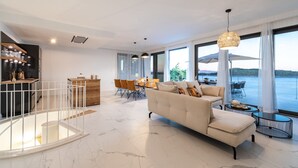 A spacious living area with a sofa, dining table, and chairs, large windows with a view of the water, and a kitchen area with a bar. - Villa Desire (Blato)