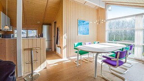 Una cocina moderna con paredes de madera, una zona de comedor con una mesa redonda y sillas coloridas, y un ventanal que ofrece vistas a árboles.