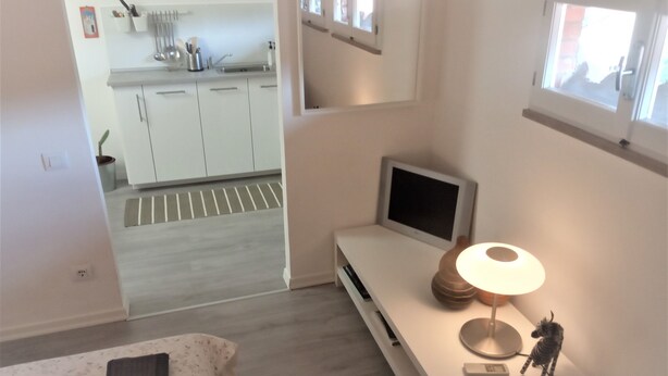 Ein modernes Hotelzimmer mit Bett, Nachttisch mit Lampe, Fernseher und Blick zur Küche.