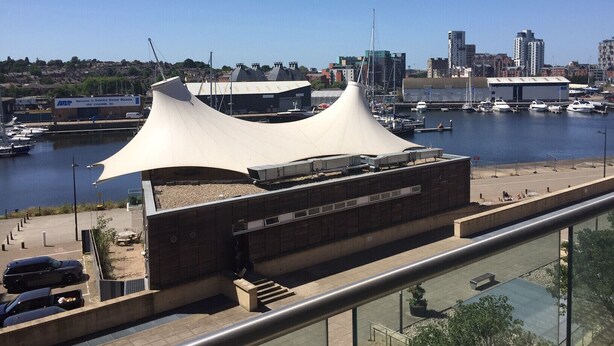 Un edifici modern amb un disseny de teulada corba i singular, ubicat a prop d'una marina amb embarcacions amarrades.