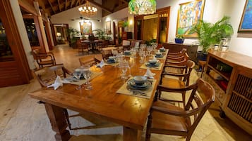 Una lunga tavola da pranzo in legno apparecchiata per un pasto, circondata da sedie in legno, con un lampadario sopra e un quadro al muro.