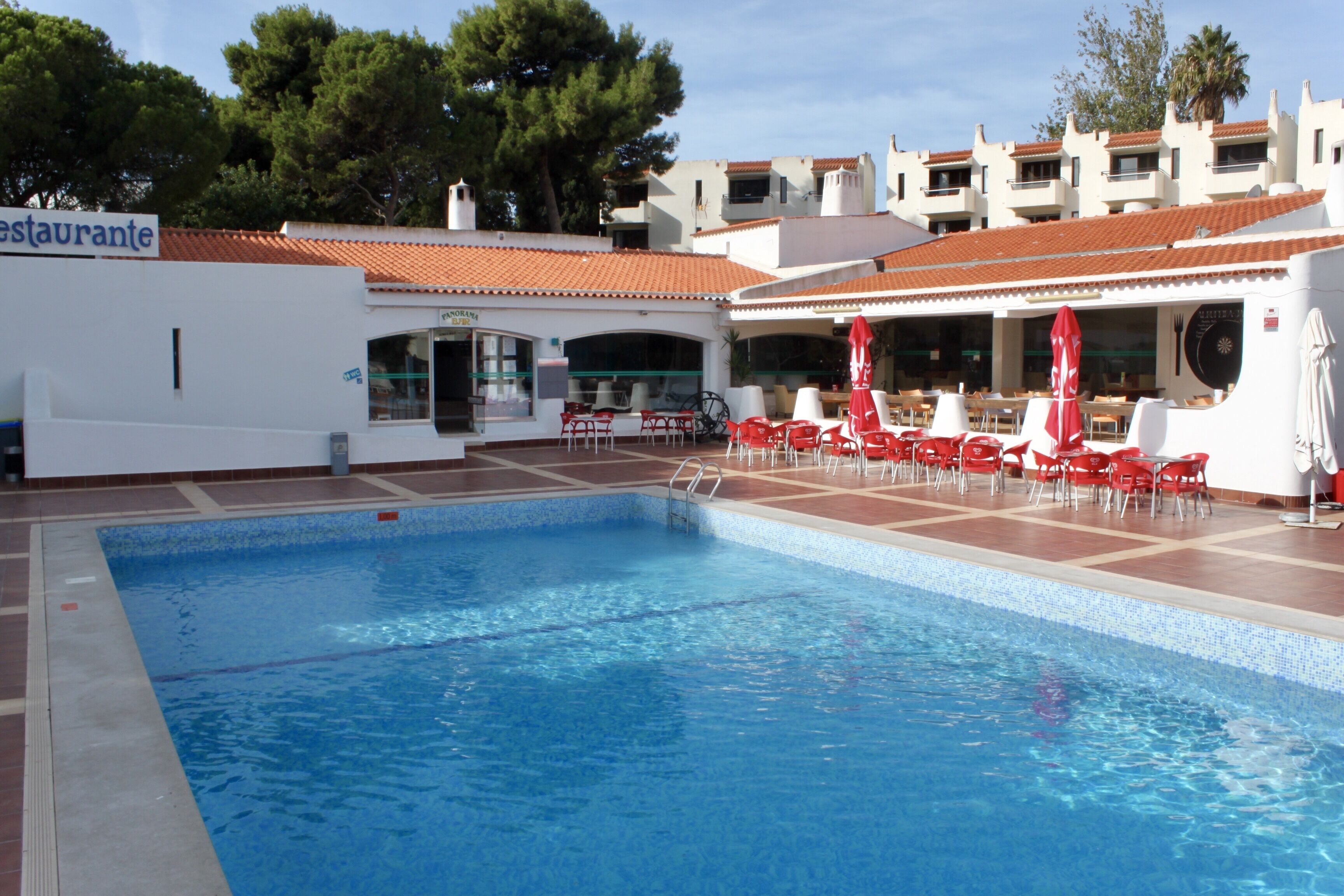 Une piscine intérieure avec des chaises rouges et des parasols, entourée d’un bâtiment doté d’un toit en tuiles et d’une enseigne de restaurant.