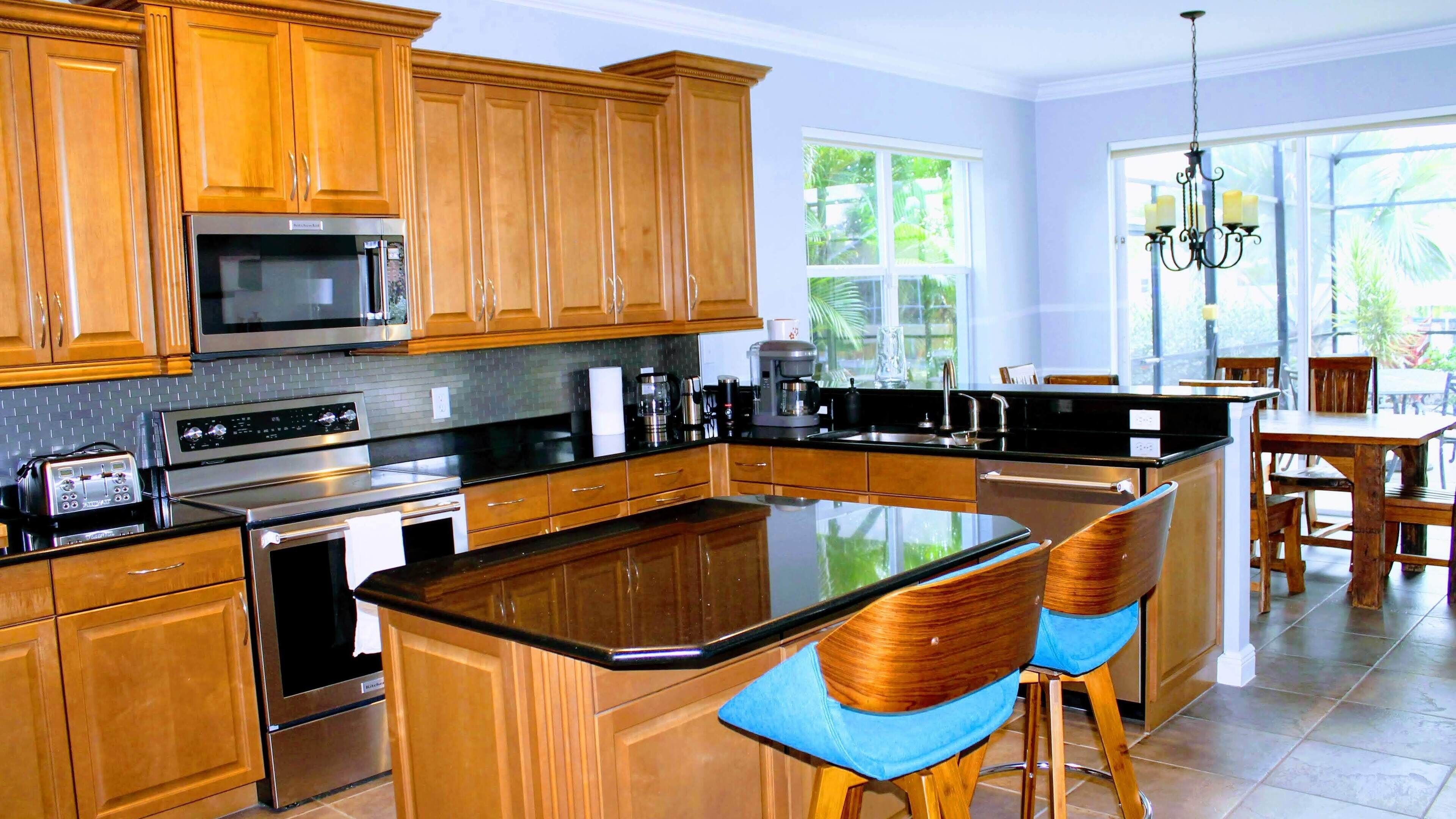 A modern kitchen with wooden cabinets, stainless steel appliances, a central island with bar stools, and large windows providing natural light.