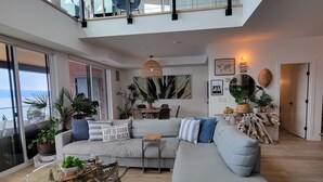 A spacious living room with a large grey sectional sofa, a glass coffee table, and a zebra-patterned rug. There are multiple potted plants, a wooden side table, and a large window offering a view of the outdoors. - EXCLUSIVE LAKEFRONT PENTHOUSE *PRIVATE ROOFTOP DECK * POOL *HOTUB *SUNSET VIEWS  (Kelowna)