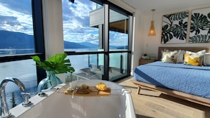 A modern bedroom with a large window, a bed with blue bedding, a bedside table with a lamp, and a view of a lake and mountains. - EXCLUSIVE LAKEFRONT PENTHOUSE *PRIVATE ROOFTOP DECK * POOL *HOTUB *SUNSET VIEWS  (Kelowna)