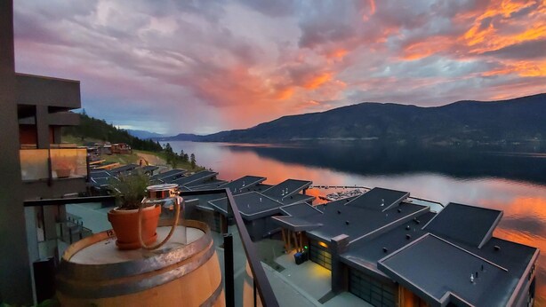 A view from a balcony overlooking a lake with mountains in the distance, modern buildings with flat roofs, and a few potted plants on the railing. - EXCLUSIVE LAKEFRONT PENTHOUSE *PRIVATE ROOFTOP DECK * POOL *HOTUB *SUNSET VIEWS  (Kelowna)