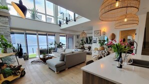 A modern living room with a large sofa, a dining area, and a kitchen with a white countertop. There are large windows and a view of the outdoors. - EXCLUSIVE LAKEFRONT PENTHOUSE *PRIVATE ROOFTOP DECK * POOL *HOTUB *SUNSET VIEWS  (Kelowna)