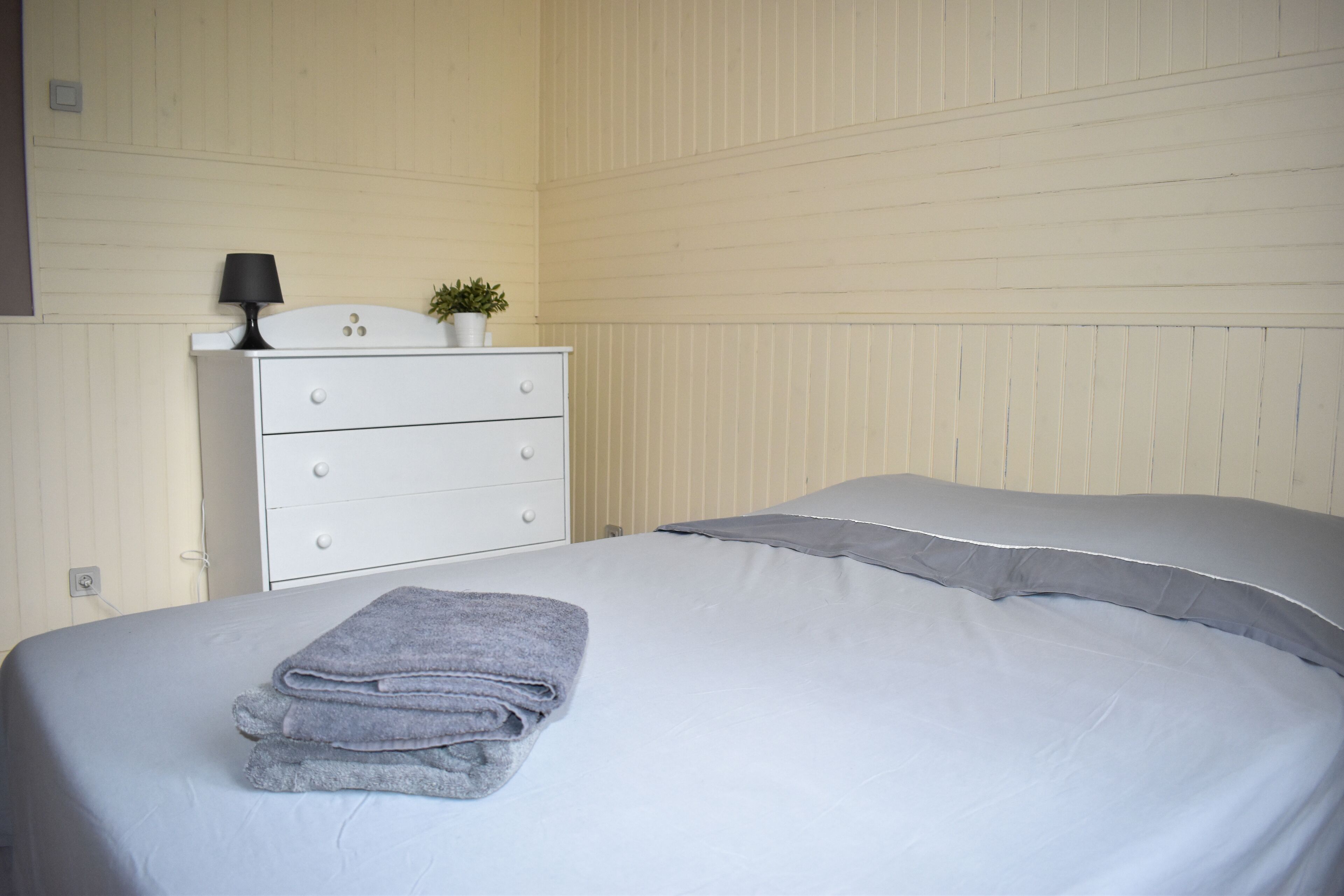 Ein ordentlich gemachtes Bett mit grauer Decke und Kissen, eine weiße Kommode mit Lampe und Pflanze sowie eine Wand mit vertikalen Linien.