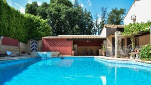 Una piscina amb una terrassa enrajolada, envoltada per un jardí i un edifici amb teulada enrajolada.