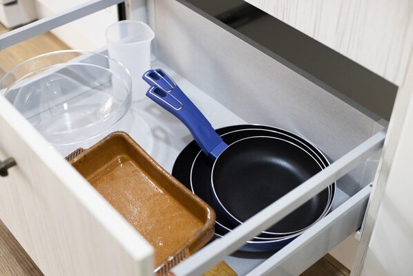 A kitchen drawer containing a frying pan, a blue spatula, a glass container, and a brown baking dish.