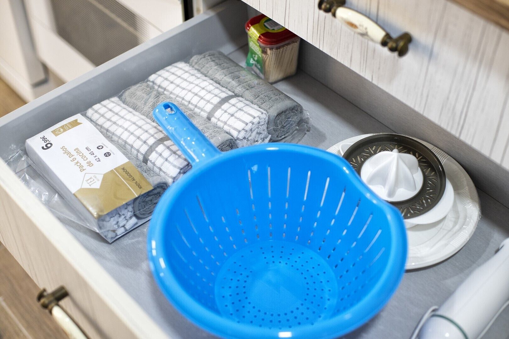 Un plan de travail de cuisine avec une passoire bleue, une brosse à vaisselle et des lingettes nettoyantes emballées.
