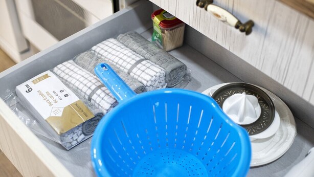 Un plan de travail de cuisine avec une passoire bleue, une brosse à vaisselle et des lingettes nettoyantes emballées.