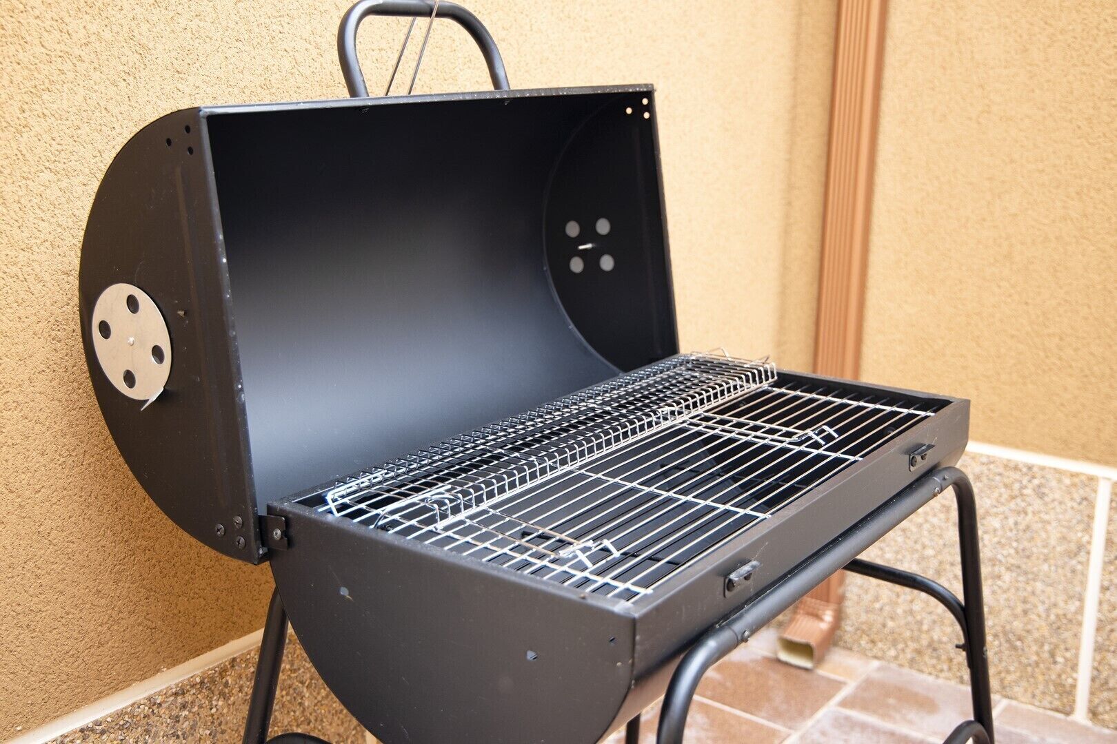 Un barbecue noir, doté d’une grille métallique, est placé contre un mur beige dont le sol est recouvert de carreaux.