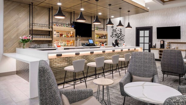 A modern bar area with a long counter, multiple seating options including bar stools and round tables, and pendant lighting.