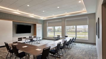 A conference room with a long table, chairs, a flat-screen TV, and large windows offering a view of the outdoors.