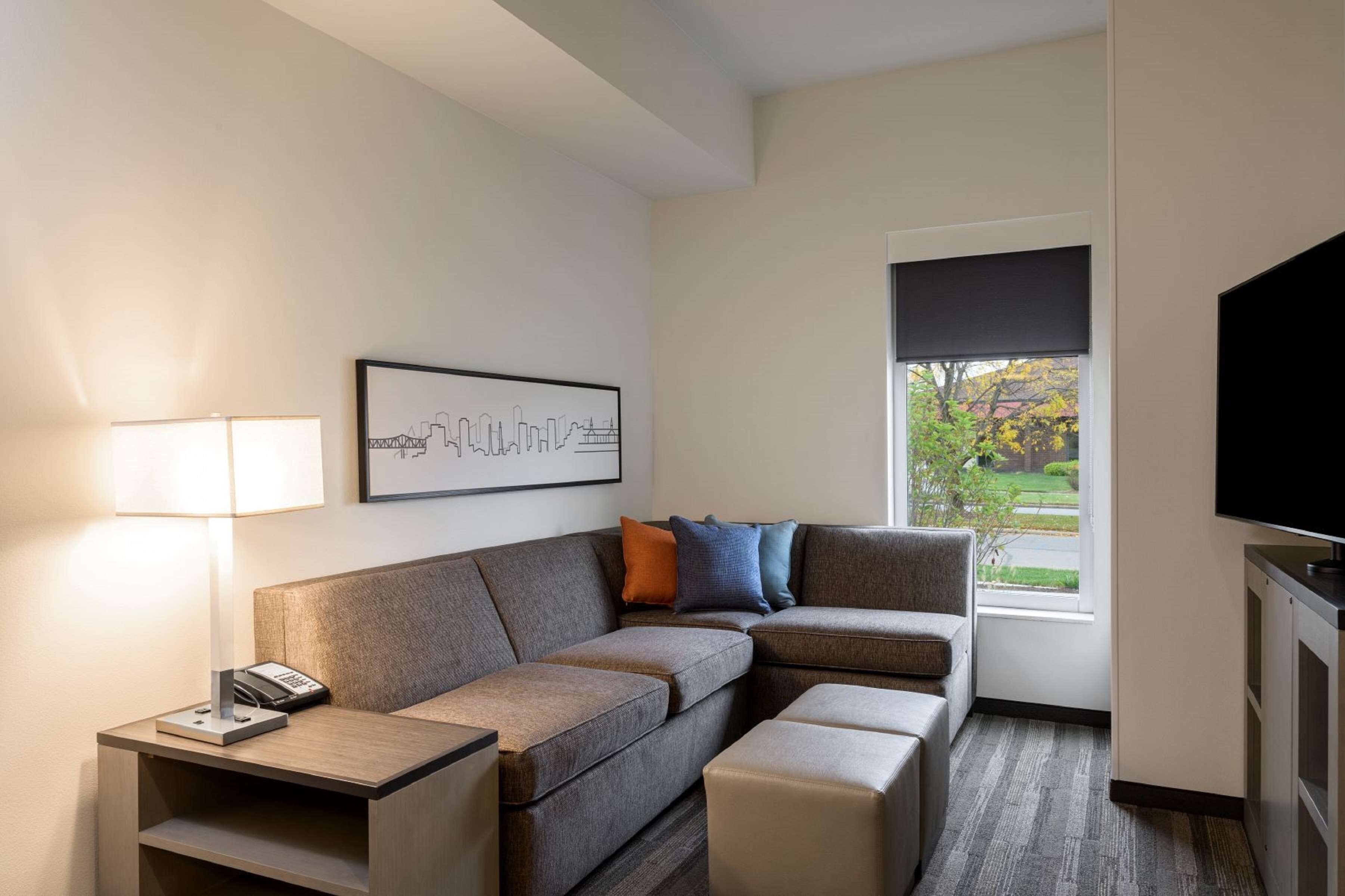 A modern hotel room with a sectional sofa, a small table with a lamp, a framed cityscape picture, a television, and a window with a view of greenery.