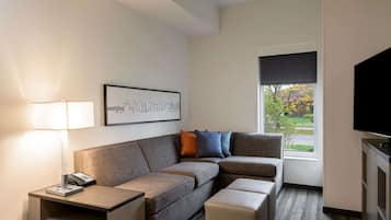 Ein modernes Hotelzimmer mit einer Eckcouch, einem kleinen Tisch mit Lampe, einem Stadtbild im Rahmen, einem Fernseher und einem Fenster mit Blick auf Grünflächen.