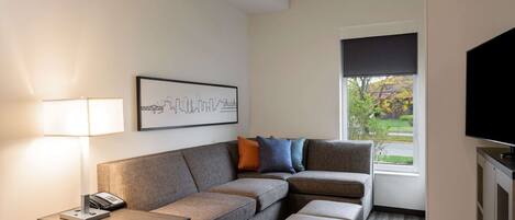 A modern hotel room with a sectional sofa, a small table with a lamp, a framed cityscape picture, a television, and a window with a view of greenery.