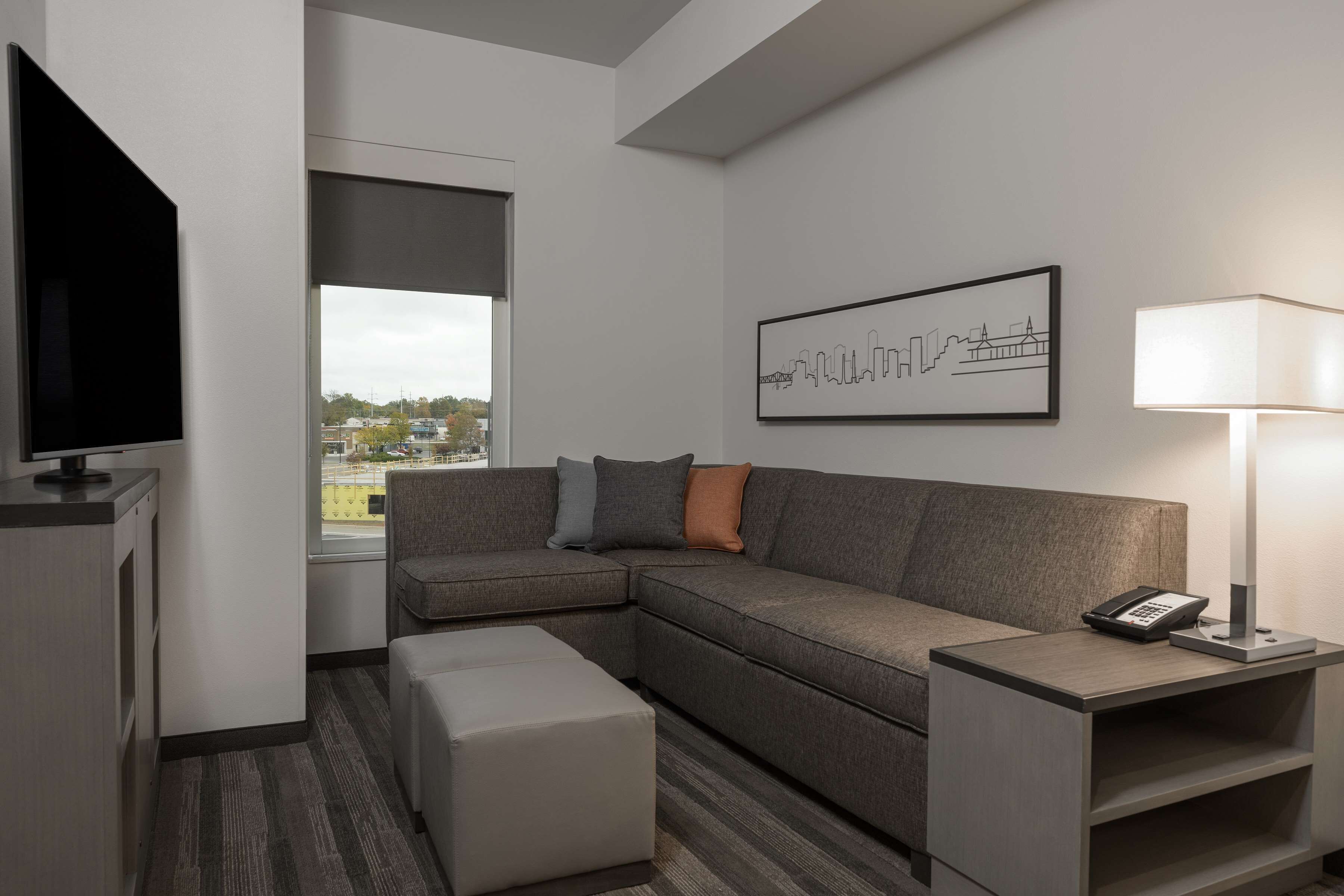 A modern hotel room with a sectional sofa, a flat-screen TV, a bedside table with a lamp, and a cityscape artwork on the wall.