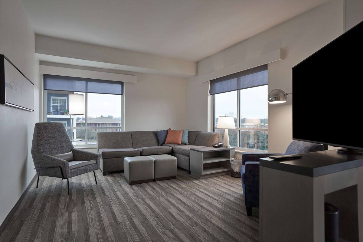 Une chambre d’hôtel moderne dotée d’un canapé d’angle, d’une télévision à écran plat et de grandes fenêtres offrant une vue sur la ville.