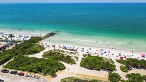 An aerial view of a beach with clear turquoise waters, a sandy shore, and numerous beach umbrellas. - Putting green, Rooftop Deck AND Beach/Gulf Views with Private Heated Pool/Spa. (Lido Key)