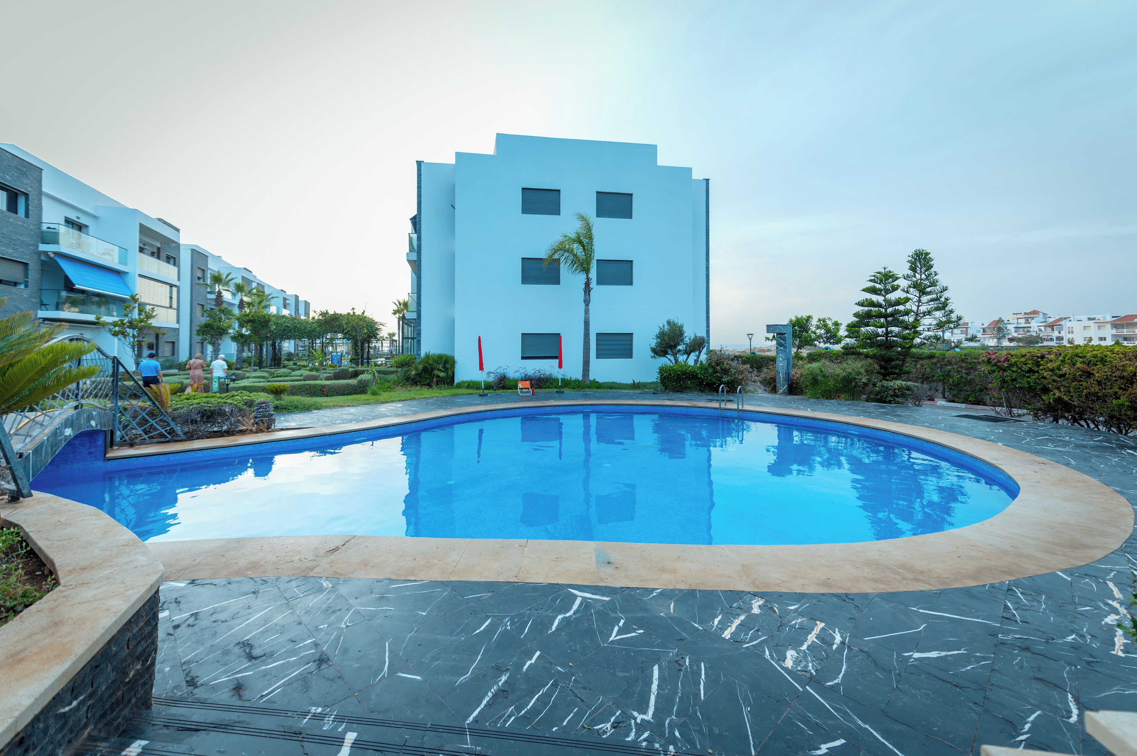 A modern hotel with a swimming pool, a well-maintained garden, and a clear view of the surrounding buildings.