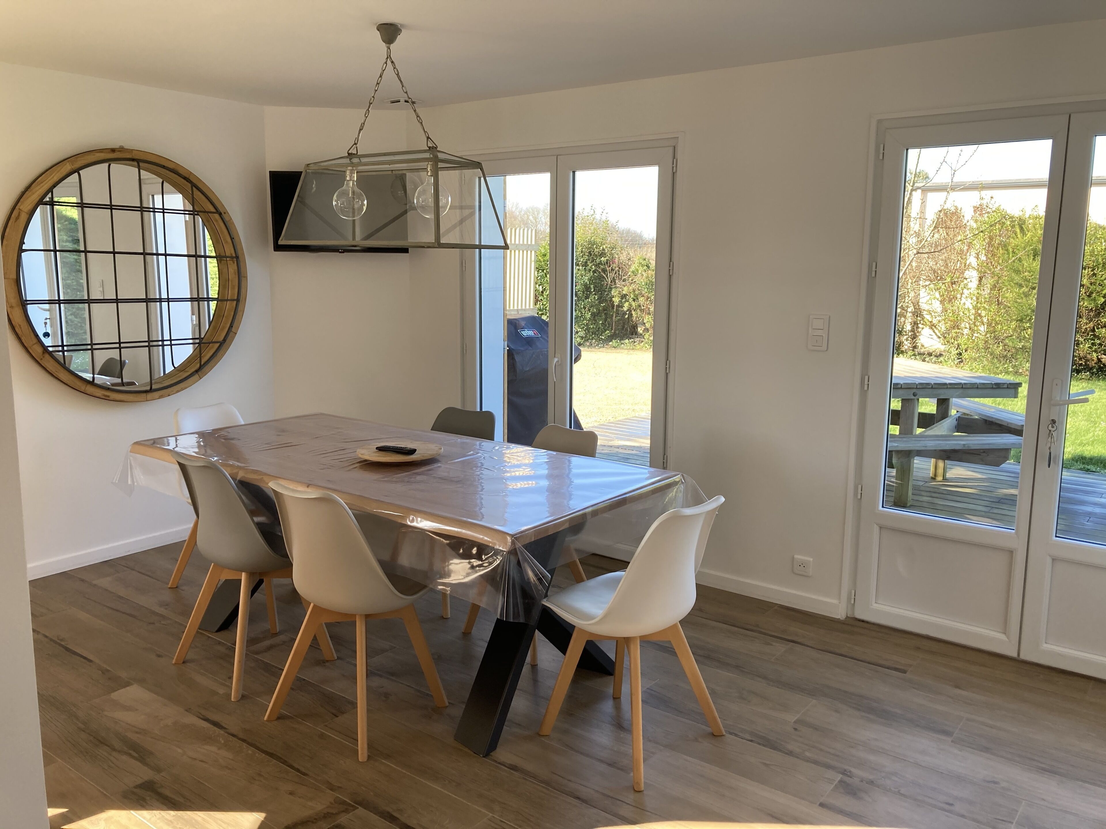 A dining area with a wooden table, white chairs, a round mirror, and a hanging light fixture.