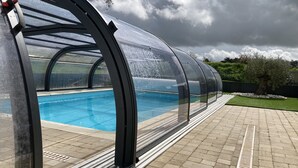 A covered swimming pool with a clear roof, surrounded by a paved patio and a grassy area.