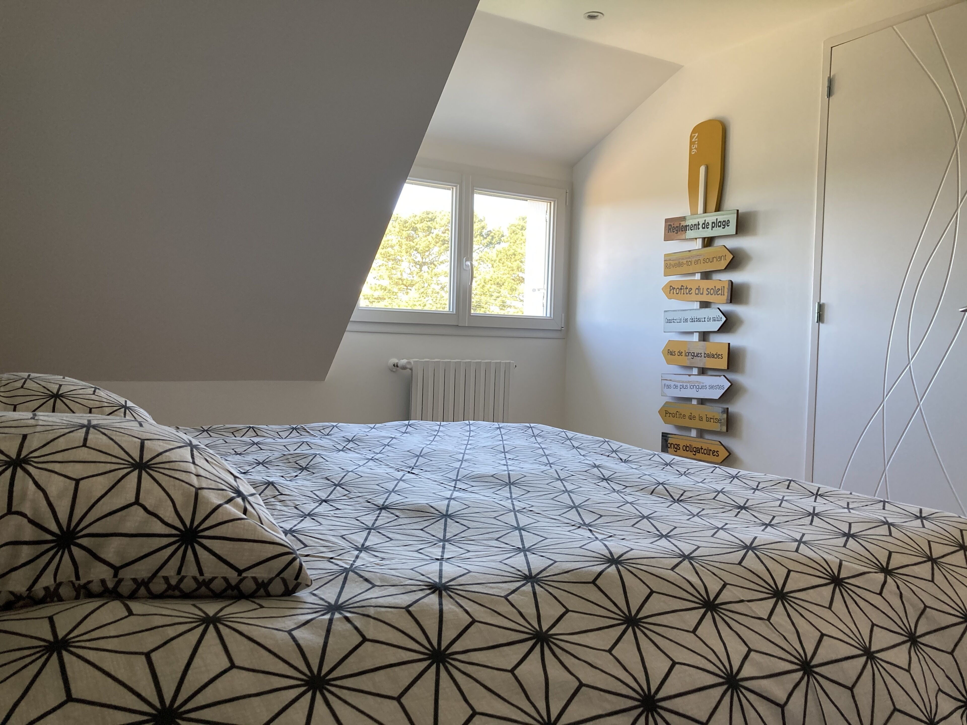 A bedroom with a sloped ceiling, a bed with patterned bedding, a window with a view of trees, and a wall with directional signs.