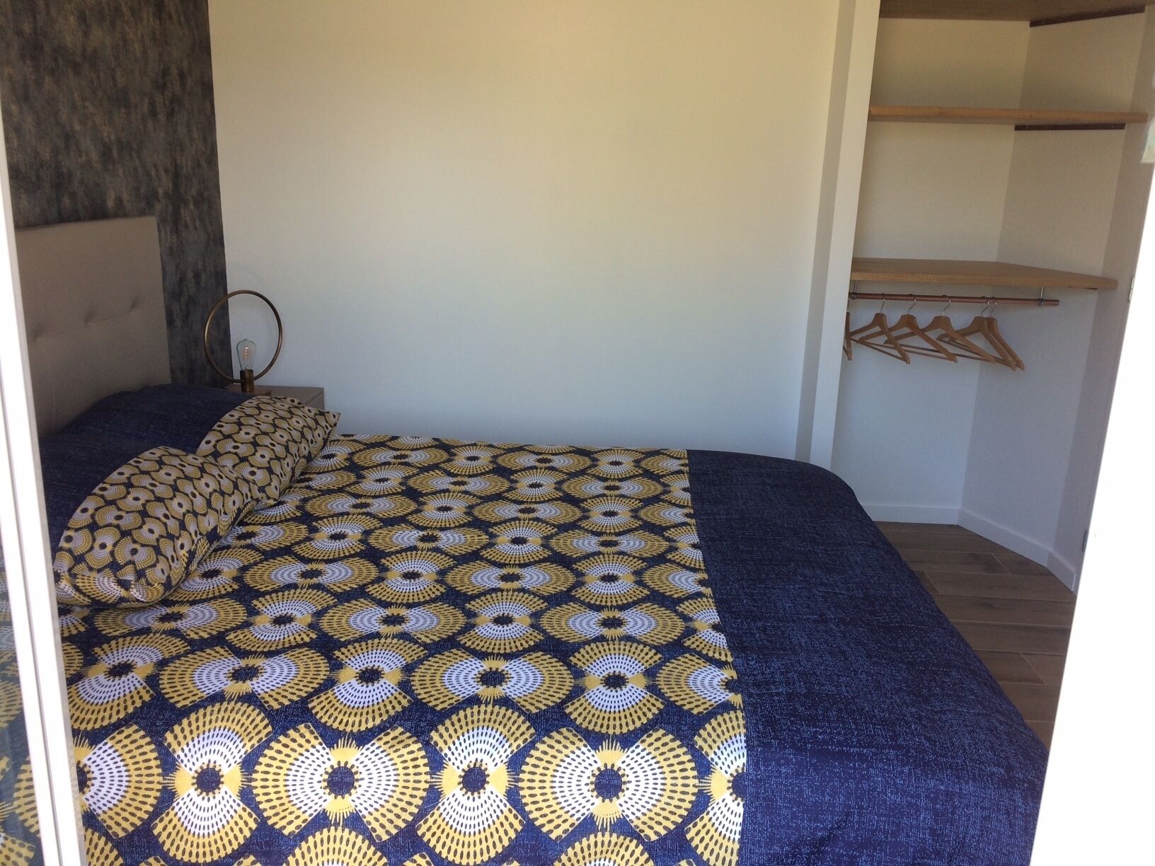 A bedroom with a patterned bedspread, a bedside table with a lamp, and a wooden shelf with hangers.