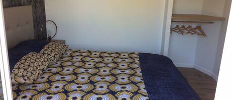 Une chambre à coucher avec une couvre-lit à motifs, une table de chevet avec une lampe et une étagère en bois avec des cintres.