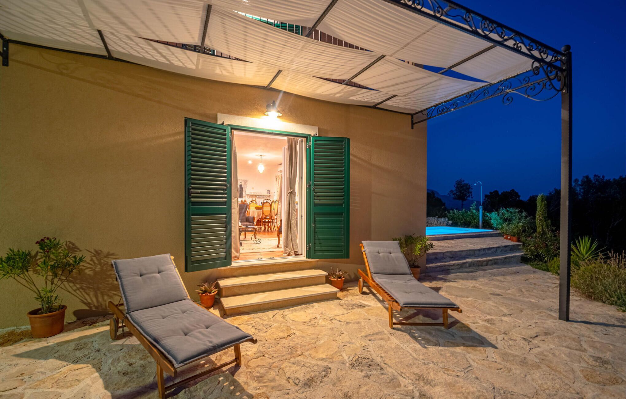 A covered outdoor seating area with two lounge chairs, a staircase leading to a door, and a pool in the background.