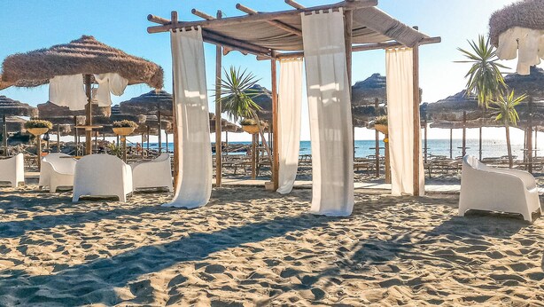 Ein Strand mit einer Holzpergola, weißen Liegestühlen und reetgedeckten Sonnenschirmen.