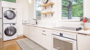 Eine moderne Küche mit eingebauter Waschmaschine, Mikrowelle und Backofen, Holzregalen und einem großen Fenster mit Blick auf Bäume.