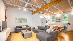Ein modernes Wohnzimmer mit grauen Sofas, einem Couchtisch und einem Fernseher. Es gibt eine Küchenzeile mit einem Esstisch und Stühlen.