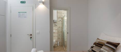 A bedroom with a bed, a pillow, a towel, a door with an exit sign, and a bathroom visible through an open door.