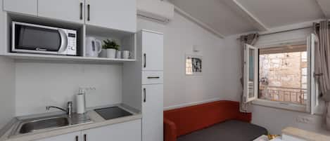 A compact kitchenette with a microwave, kettle, and sink. There is a small seating area with a red cushion and a window with curtains.