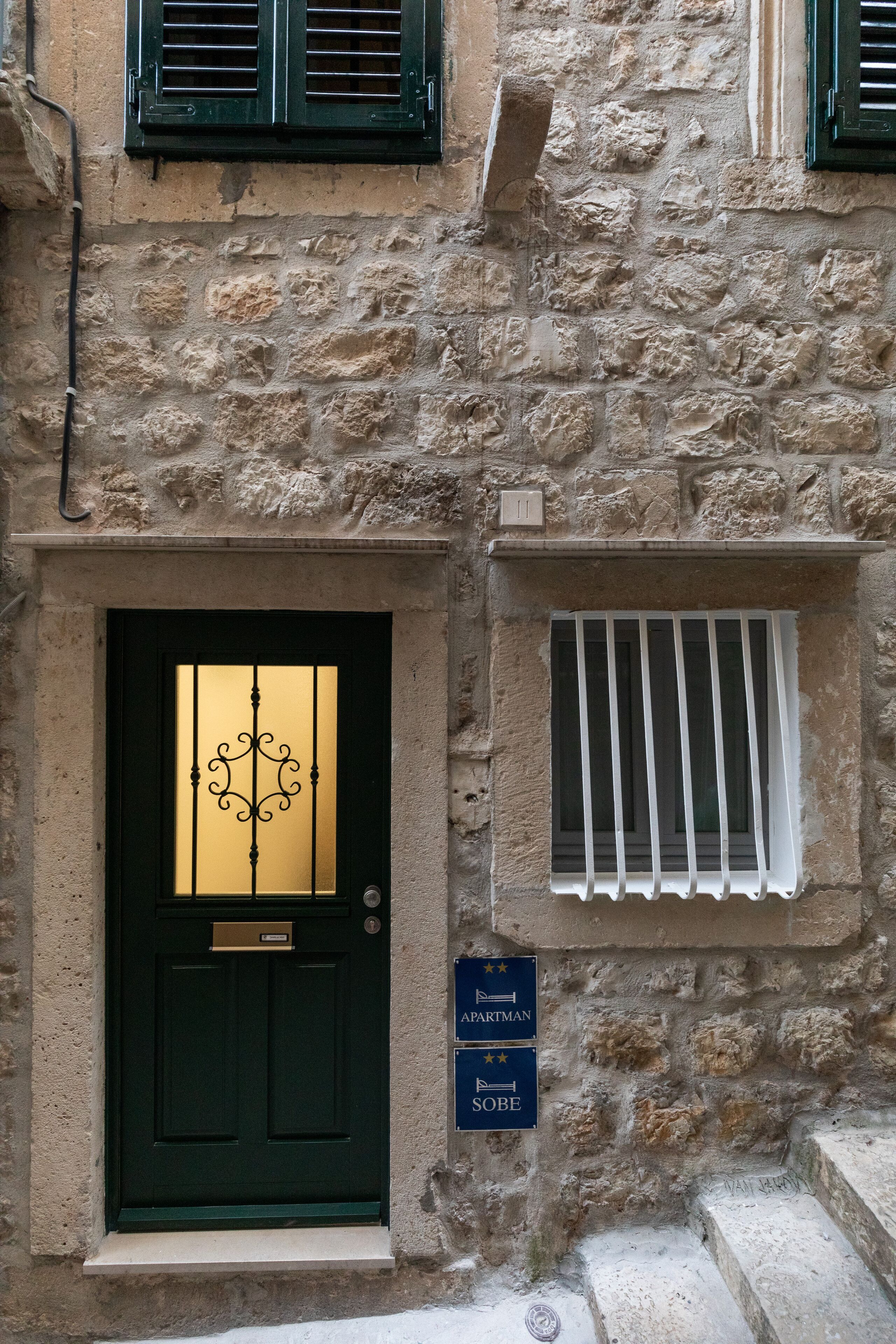 Ein Steinhaus mit einer schwarzen Tür und einem vergitterten Fenster. Schilder weisen 'APARTMAN' und 'SOBE' aus.