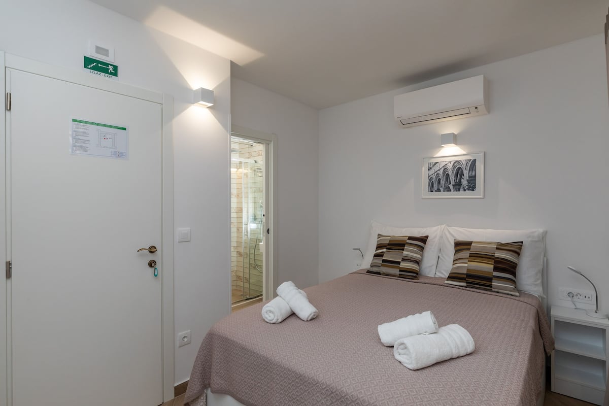Hotelkamer met een bed, twee kussens, een nachtkastje, een airconditioning en een deur met een bordje.