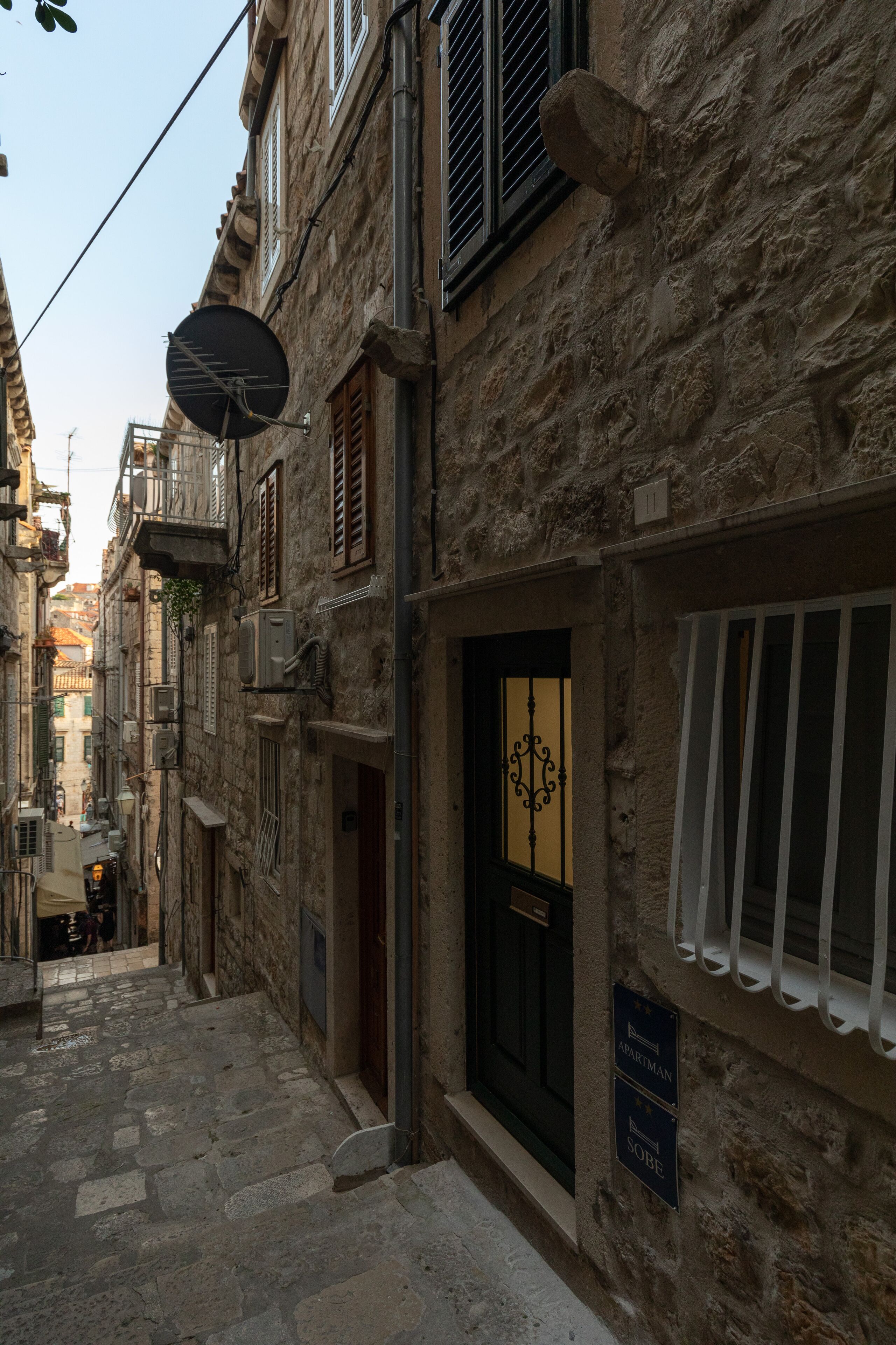 A narrow street with stone buildings, a visible door with a sign, and a satellite dish.