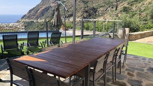 Eine Terrasse mit Holztisch und -stühlen, ein überdachter Sitzbereich und Blick auf das Meer und die Berge.