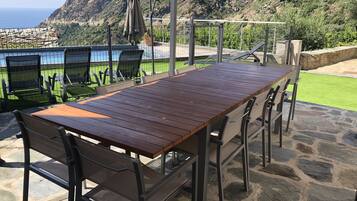 Un espace patio avec une table et des chaises en bois, un coin salon abrité et une vue sur l’océan et les montagnes.