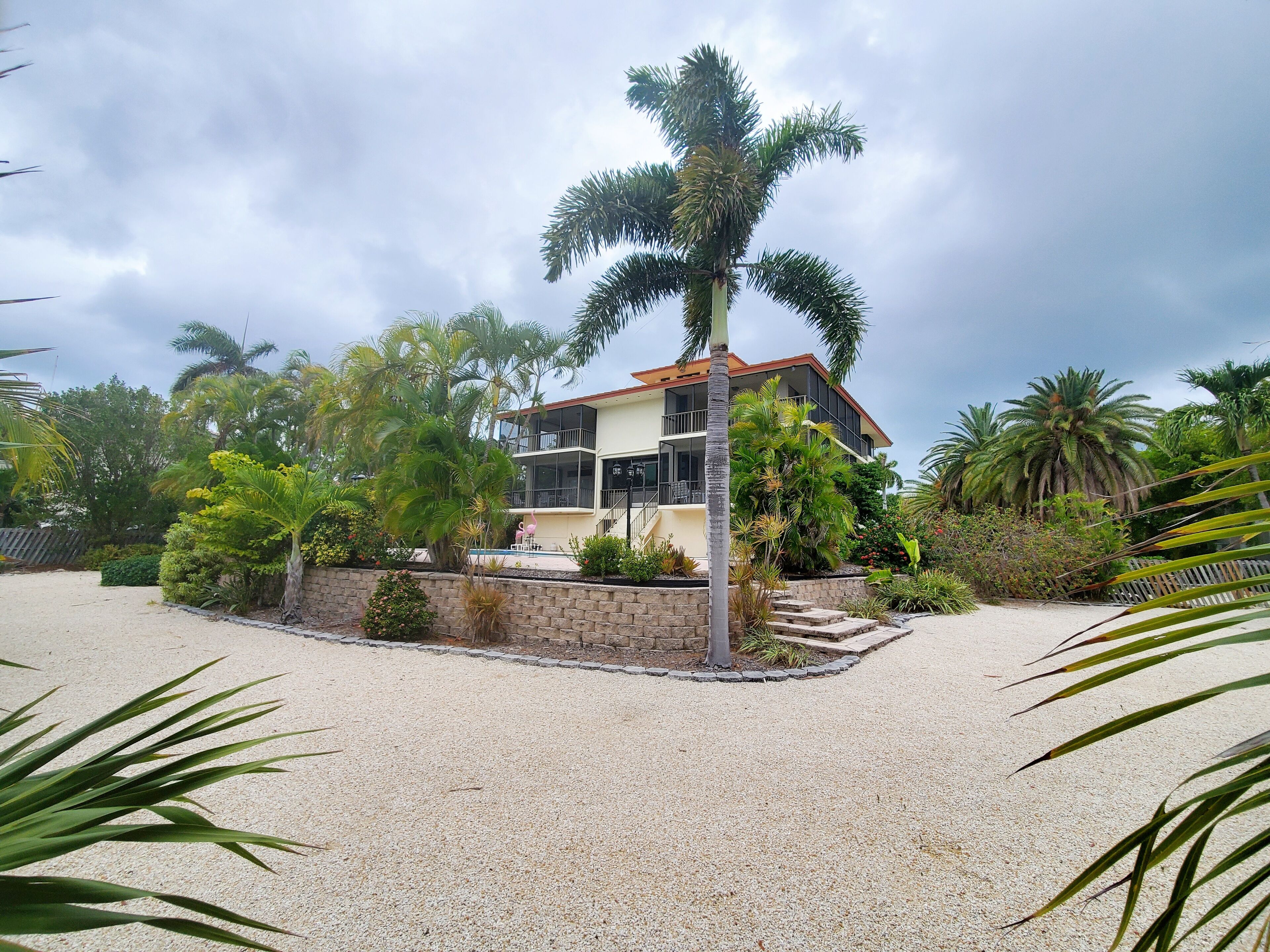 A two-story house with a balcony, surrounded by palm trees and a gravel driveway.