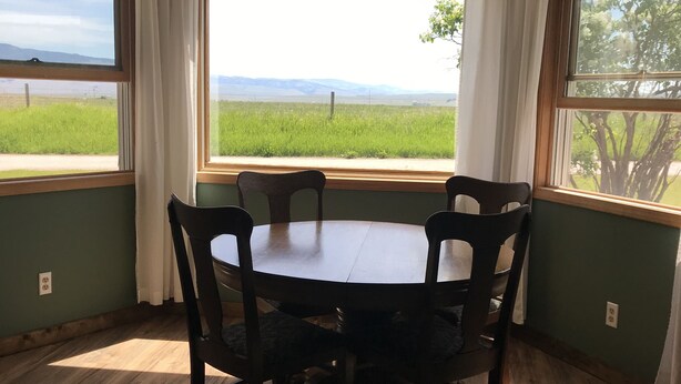 Ein Esszimmer mit Tisch und Stühlen, großen Fenstern mit Vorhängen und Blick auf ein grünes Feld und Berge.